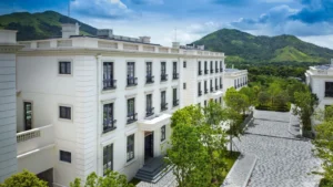 White multi story residential building with balconies trees and a cobblestone courtyard set against green hills behind it