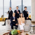 Five business professionals in a bright office lobby pose for a group photo two seated on a sofa and three standing giving thumbs up with a city skyline visible through large windows