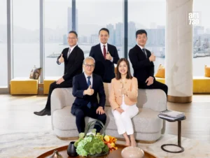 Five business professionals in a bright office lobby pose for a group photo two seated on a sofa and three standing giving thumbs up with a city skyline visible through large windows