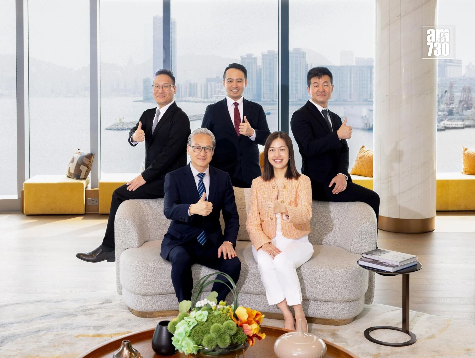 Five business professionals in a bright office lobby pose for a group photo, two seated on a sofa and three standing, giving thumbs up with a city skyline visible through large windows.