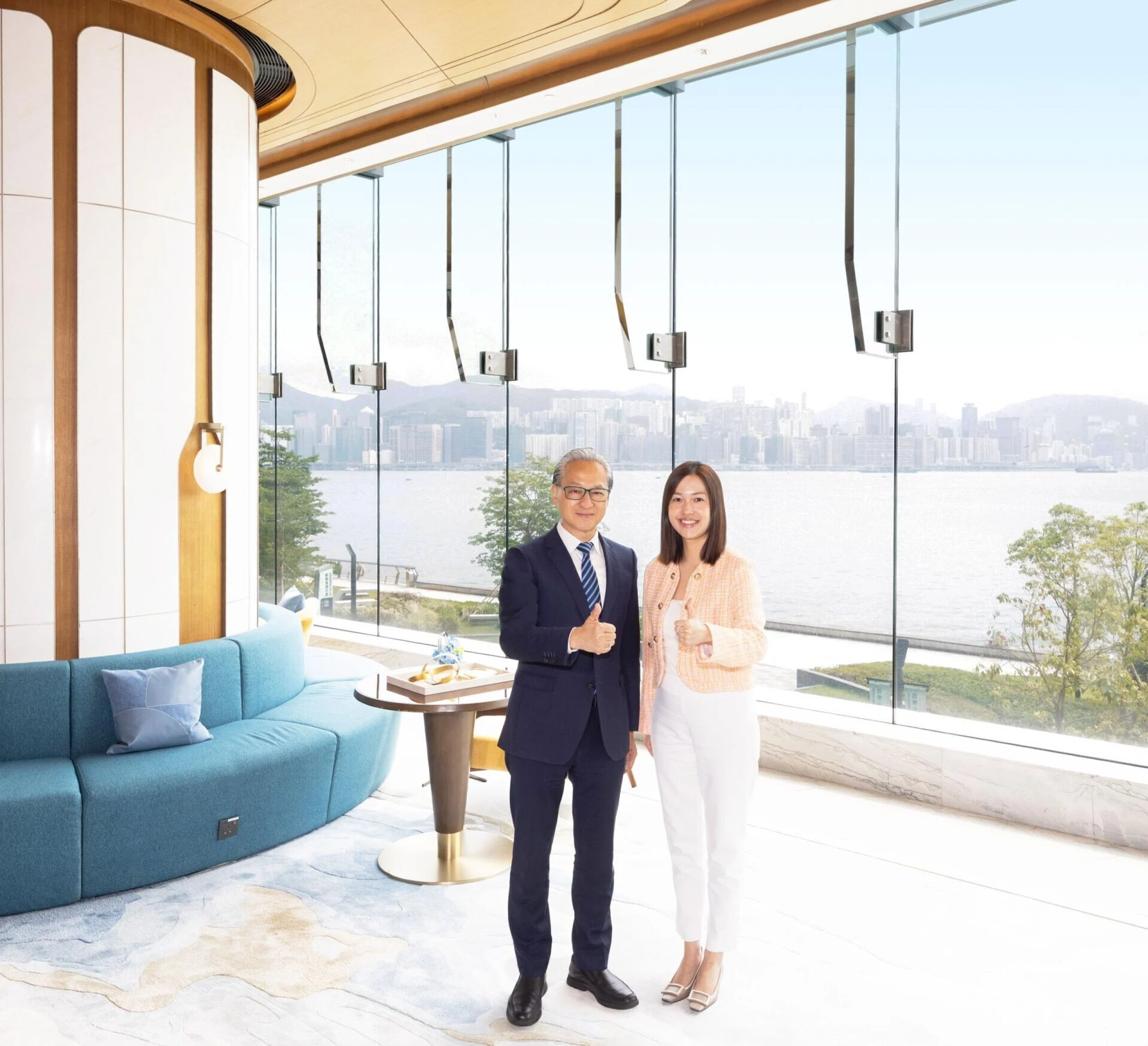 Two professionals in a bright glass-walled lounge pose and give thumbs up with a city skyline and water in the background.