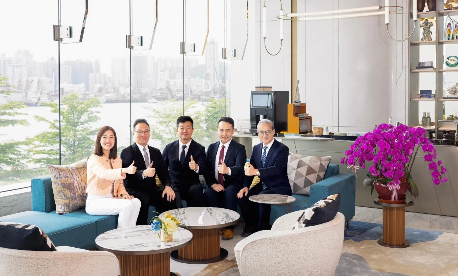 Five professionals in business attire sit on a blue sectional in a bright modern lounge, giving thumbs up with a city skyline visible through glass walls and purple orchids on the right.