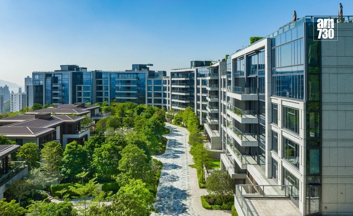 Modern residential complex with glass-front buildings, a tree-lined pedestrian path, and landscaped gardens along a central walkway. A logo is visible in the top-right corner.