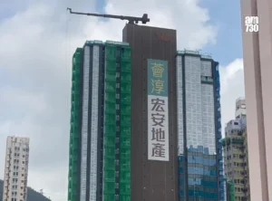 Under construction high rise with green safety netting and a vertical banner of Chinese text crane arm at the top and an am730 logo in the corner