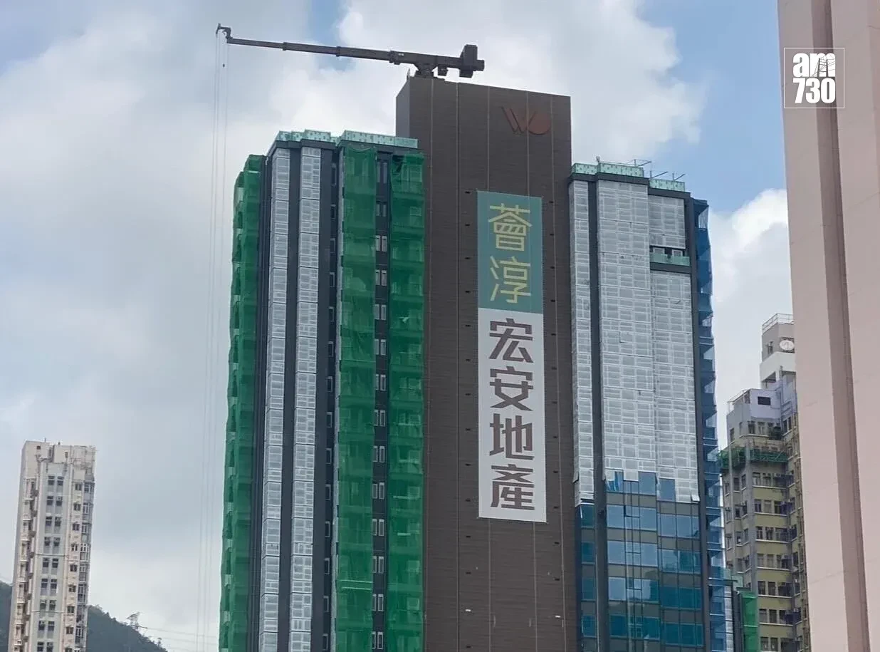 Under-construction high-rise with green safety netting and a vertical banner of Chinese text; crane arm at the top and an am730 logo in the corner.