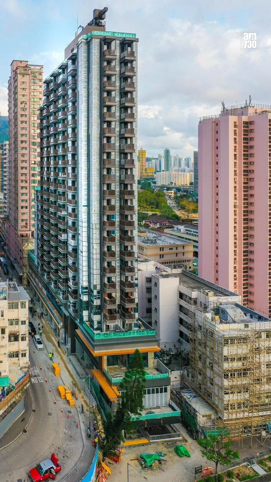 Tall modern high-rise under construction in a dense city, with a curved road and pink building to the right.