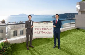 Two professionally dressed men stand on a balcony holding a large real estate poster with Chinese text overlooking the sea and distant hills with a logo in the top right corner