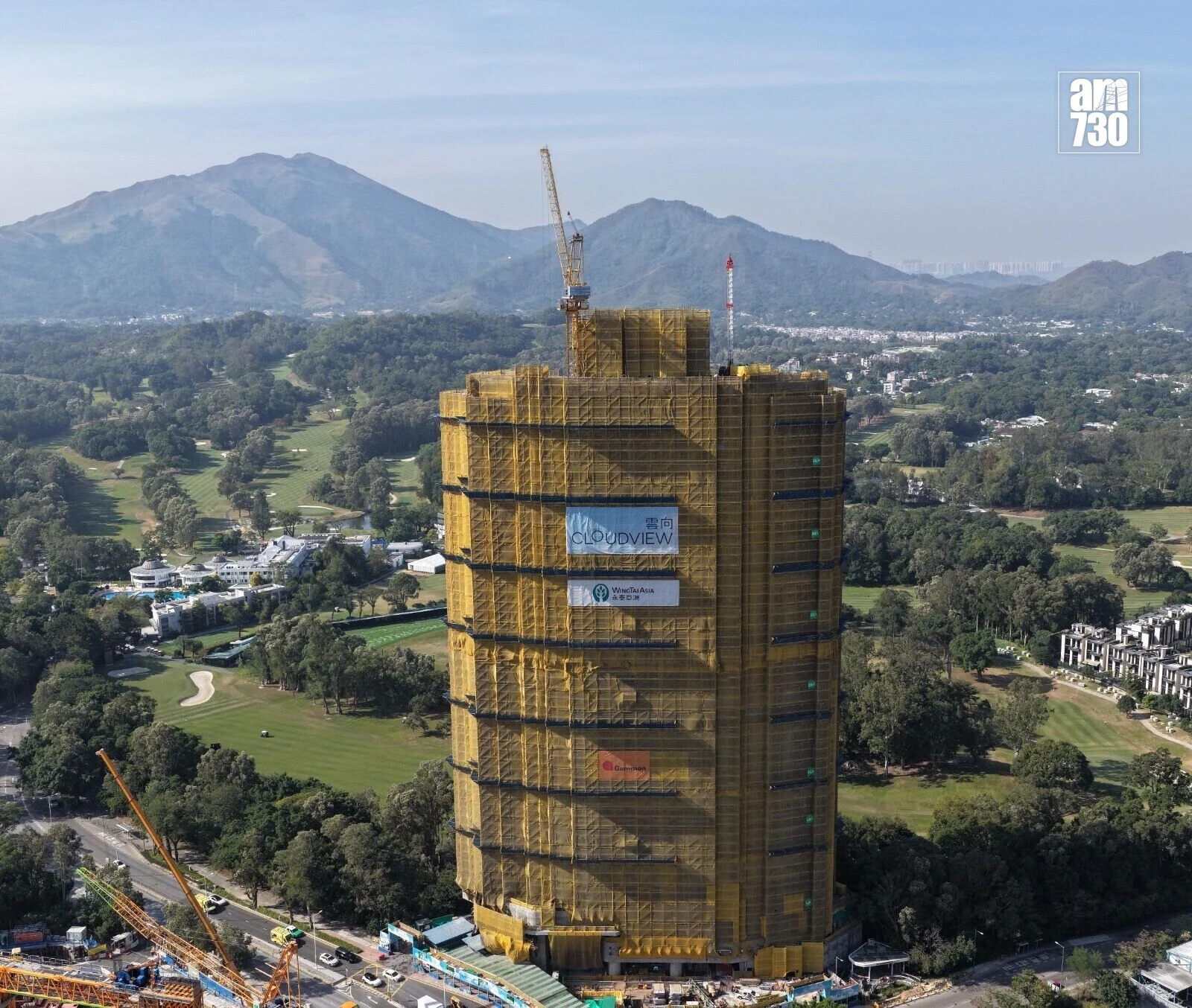 Tall high-rise under construction with a crane on top, wrapped in yellow safety scaffolding, set against green landscape and distant mountains.
