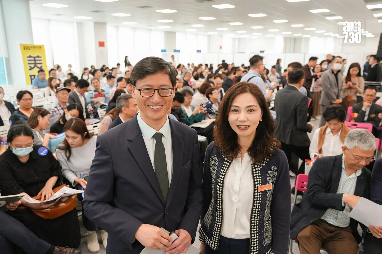 Two smiling colleagues pose for a photo at a crowded conference hall with attendees seated and mingling in the background.