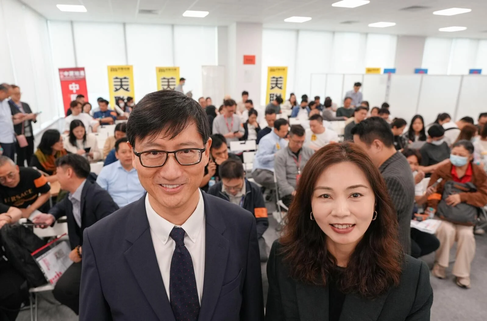 Man in a dark suit and glasses taking a selfie with a smiling woman beside him at a crowded conference hall.