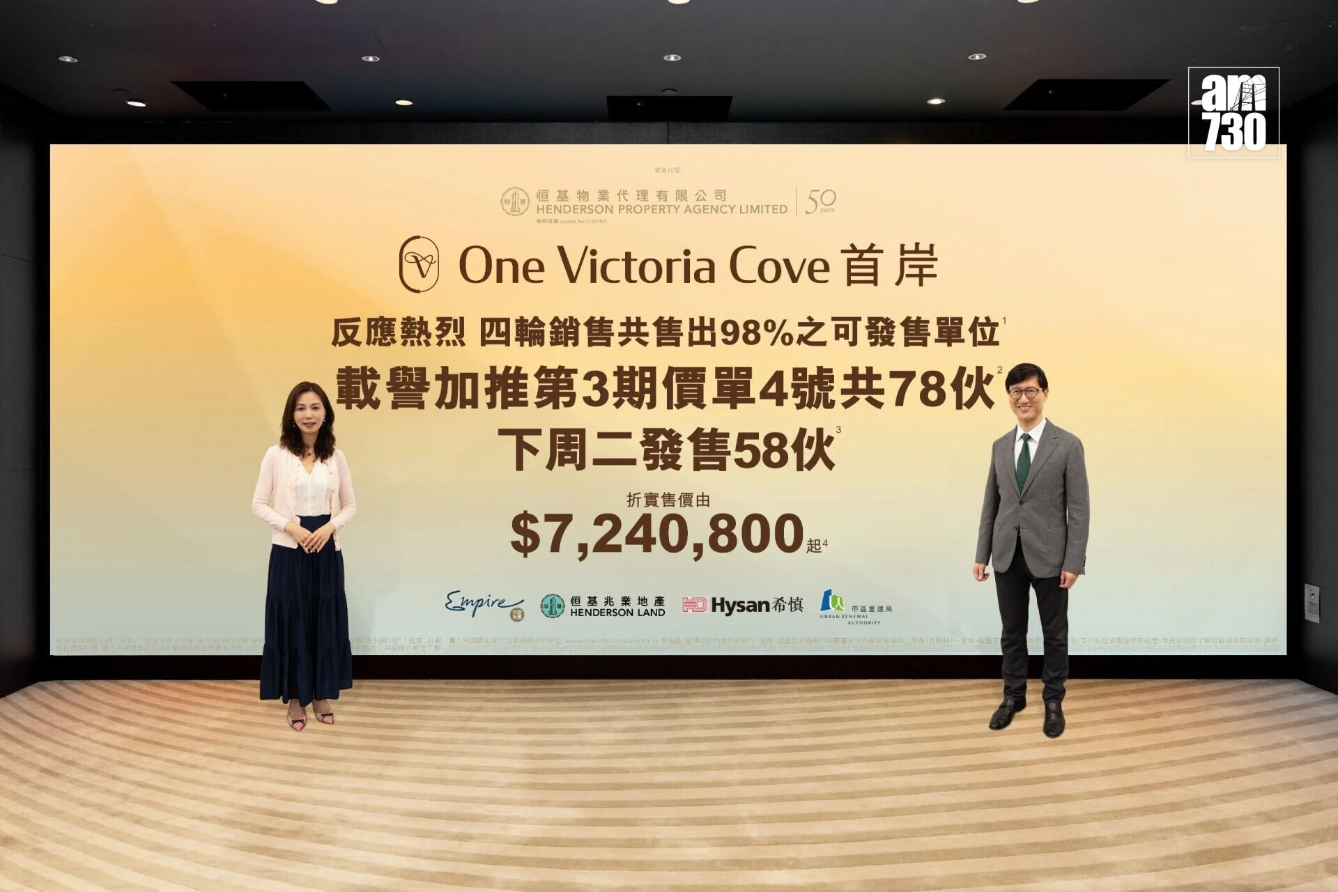 Two presenters stand in front of a large yellow promotional banner reading 'One Victoria Cove' with Chinese text and logos above, indoors on a stage.