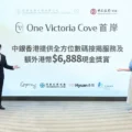 Two presenters stand on a stage in front of a large screen reading One Victoria Cove with Chinese text and sponsor logos visible