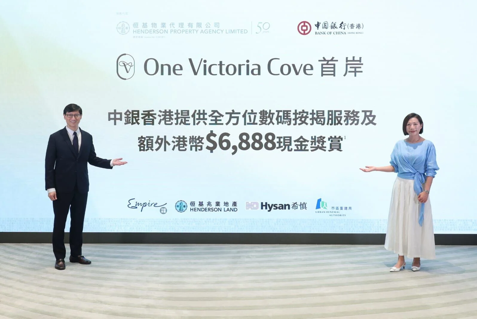 Two presenters stand on a stage in front of a large screen reading 'One Victoria Cove' with Chinese text and sponsor logos visible.