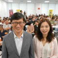 Smiling man in a gray suit and a woman in a light blouse pose at the front of a crowded conference room