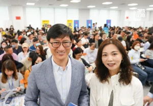 Two smiling people take a selfie at a large indoor event with hundreds of attendees in the background