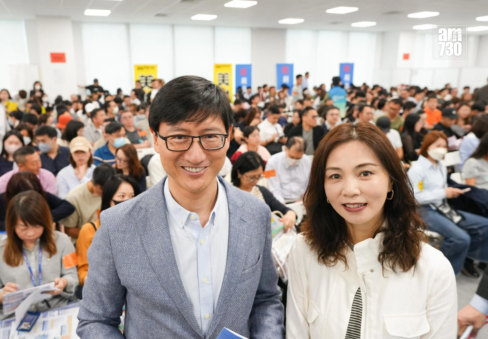 Two smiling people take a selfie at a large indoor event with hundreds of attendees in the background.
