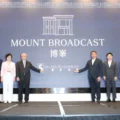 Seven professionals in formal attire stand on a stage in front of a blue backdrop reading 'MOUNT BROADCAST' with company logos, posing for a photo.