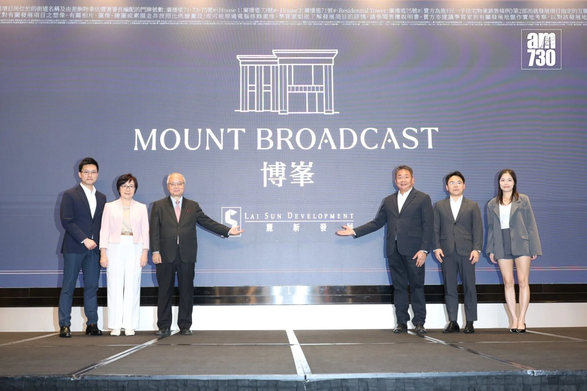 Seven professionals in formal attire stand on a stage in front of a blue backdrop reading 'MOUNT BROADCAST' with company logos, posing for a photo.